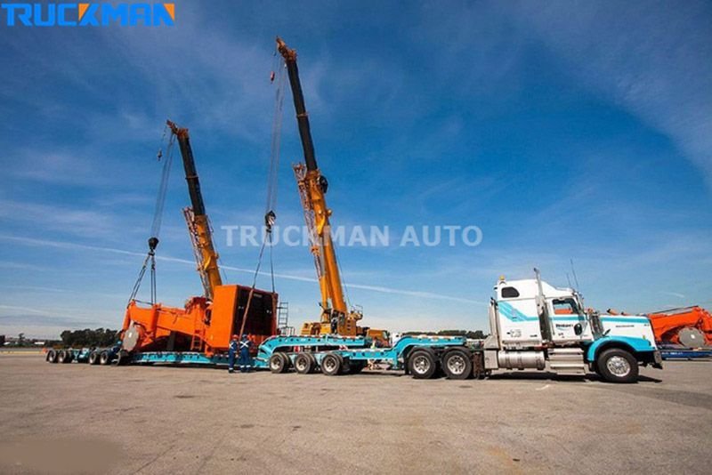 Duas gruas móveis a levantar equipamento pesado para um semirreboque de caixa baixa